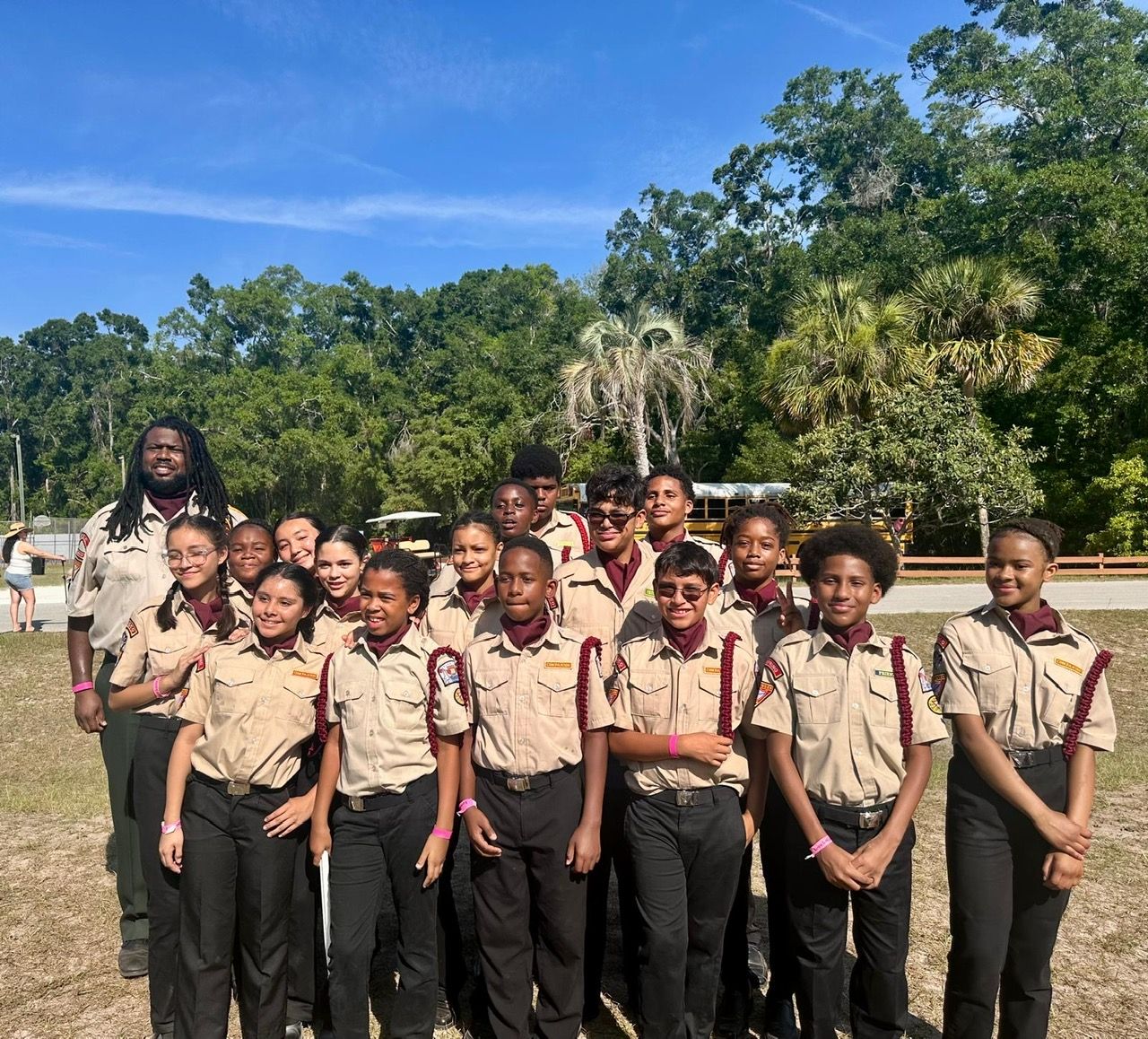 Drum corps members and an instructor smiling together outdoors in khaki uniforms with burgundy neckerchiefs and cords.