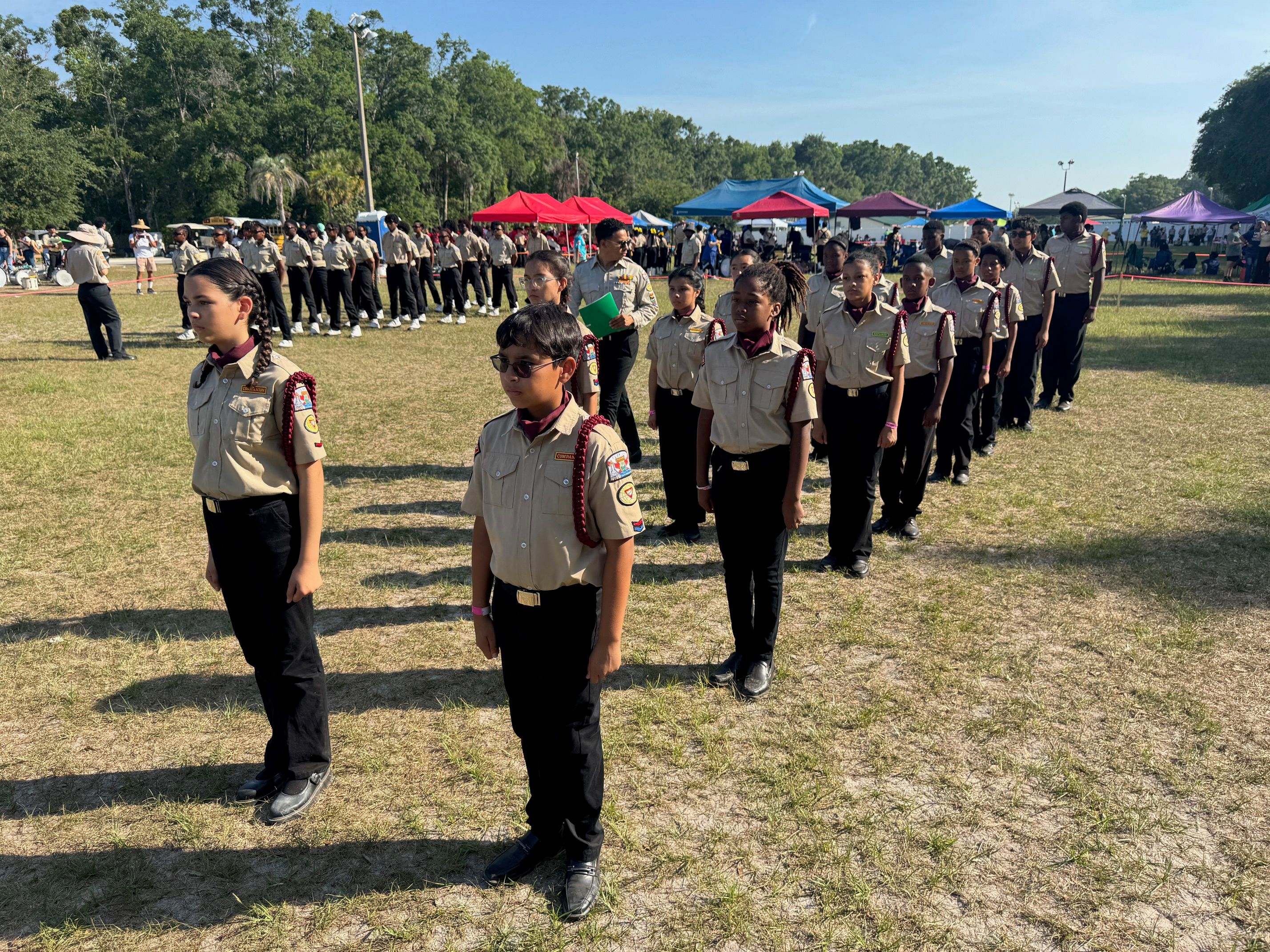 Drum corps members standing in formation on a grass field in khaki uniforms with burgundy cords.