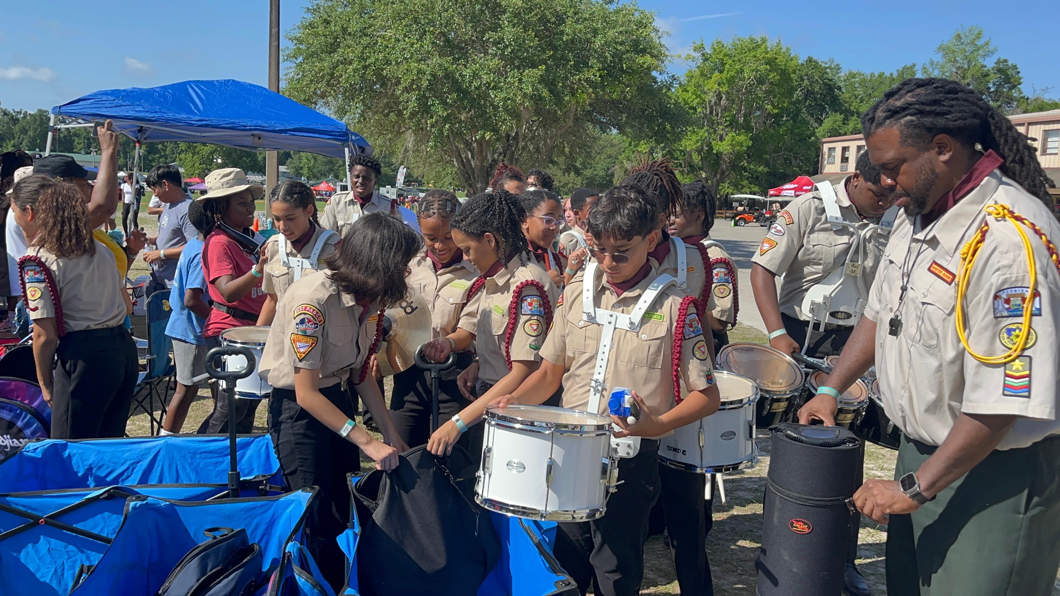 Drum corps members unpacking white snare drums at an outdoor event while an instructor helps.